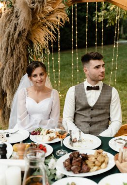 Genç ve güzel gelin ve damat kemerin arkasındaki düğün masasında oturuyorlar. Kova üzerinde yeni evliler. Düğün yemeği servisi. Kırsal bir düğün. ahşap kemer