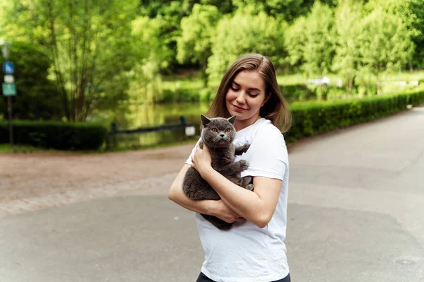 a cute girl holds a gray cat in her arms while being outside in nature. girl with a cat and a carrier. a walk with a pet in the air