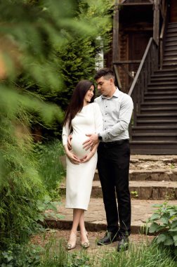 Evli bir çift, ahşap bir ekoloji evinin arka planına nazikçe sarılıyor. Karım hamile. Adam nazikçe hamile bir kadına sarılıyor.