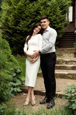 Evli bir çift, ahşap bir ekoloji evinin arka planına nazikçe sarılıyor. Karım hamile. Adam nazikçe hamile bir kadına sarılıyor.