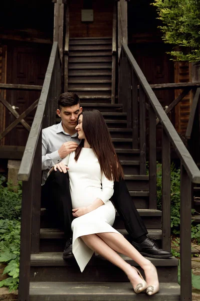 妊婦の男が家の入り口近くの木の階段に座っている 美しい妊娠中のカップル — ストック写真