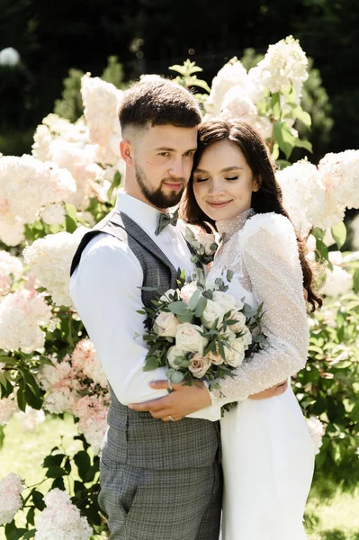 一对美丽的新婚夫妇在花朵的衬托下拥抱在一起 新娘和新郎穿着漂亮的婚纱散步 — 图库照片