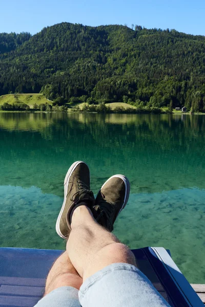 The legs of a man sitting on a deckchair, on the shore of a lake