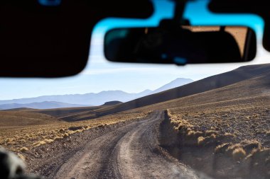 Avaroa Ulusal Parkı 'ndan geçen uzak bir toprak yolda giden bir aracın içinden görüntüyü al. Dramatik dağ siluetleri ve ufka kadar uzanan çöl manzaraları..