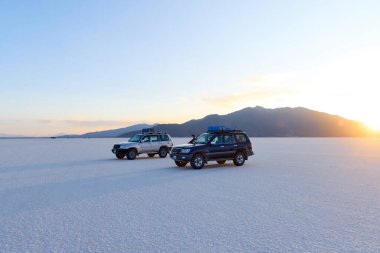 Gün batımında parlak beyaz Salar de Uyuni tuzuna park edilmiş iki 4x4 araç. Toyota Land Kruvazörleri Bolivya 'daki dağ zeminine karşı çatı ekipmanlarıyla. 16.10.2023, Bolivya