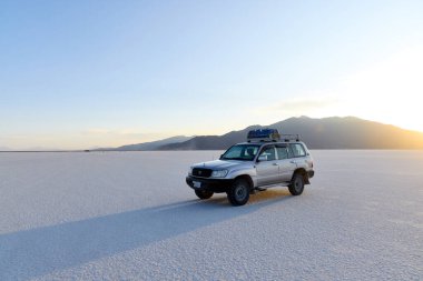Beyaz Toyota 4x4 araç gün batımında geniş Salar de Uyuni tuz düzlüğünde. Bolivya 'nın ünlü manzarasında dramatik dağ siluetleri ve el değmemiş beyaz tuz tabakası. 16.10.2023, Bolivya