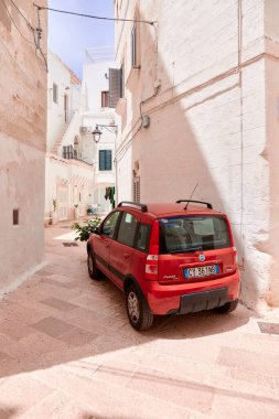 Kırmızı bir Fiat Panda 4x4 dar, beyaz bir sokağa park etmiş. Küçük şehir arabası bu eski kasabadaki geleneksel kireçtaşı binalar arasındaki dar alana mükemmel şekilde uyuyor. 25.06.2022, Monopoli, İtalya
