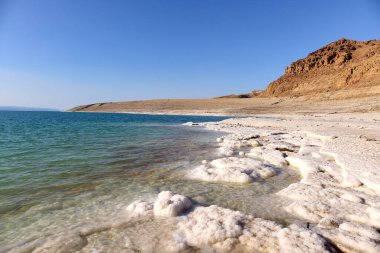 Ürdün 'deki Ölü Deniz kıyılarının manzarası. Sığ sularda tuz oluşumları var. Turkuaz sular, berrak mavi gökyüzünün altında aşınmış dağ arkaplanlarına karşı beyaz tuz yataklarıyla buluşur..