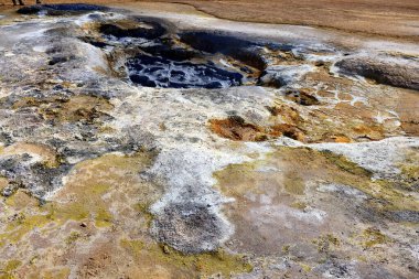 Hver, İzlanda. Canlı jeotermal özellikler, engebeli bir arazide mineraller ve köpüren çamurların bir karışımını ortaya çıkarıyor. Ziyaretçiler parlak güneş ışığı altında dinamik manzarayı gözlemliyorlar.