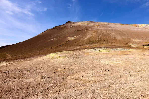 Hver, İzlanda. Issız bir tepe berrak mavi bir gökyüzüne karşı yükselir ve çıplak toprak ve kayalarla karakterize edilmiş kurak araziyi gösterir. Bu volkanik arazi uzak bir bölgedeki doğanın gücünü ortaya koyuyor..