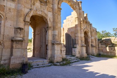 Ziyaretçiler Ürdün 'de bulunan tarihi eser Jerash' ın anıtsal kemerlerinden geçiyorlar. İyi korunmuş yapılar, açık mavi gökyüzü altında Roma mimarisini sergiliyor..