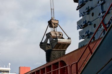 Büyük bir vinç, yoğun bir rıhtımdan bir gemiye kömür yüklüyor. Kargo bulutlu bir günde bekleyen kamyonlara boşaltılırken işçiler süreci denetliyor.