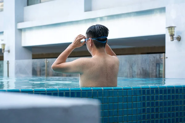 Back view of man in the pool, relaxing and adjusting his goggles by the side. Vacation or travel concept.