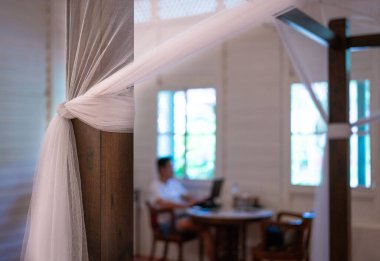 Bedroom with mosquito net tied to bedpost. Blur man working on the computer at the background. Work from home. clipart