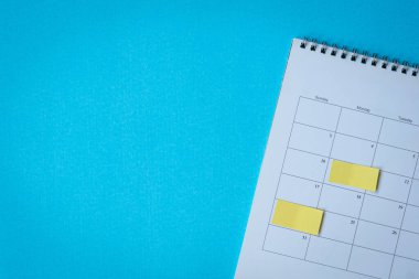 Sarı yapıştırıcılı takvim sayfası not alıyor. Mavi yüzeyin üstünde, yüksek açı manzarası. Boşluğu kopyala.