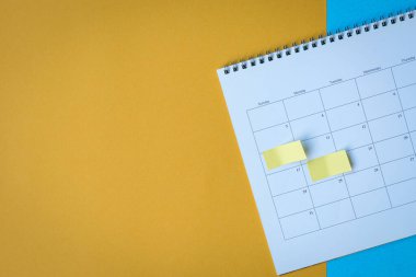 Sarı yapıştırıcılı takvim sayfası not alıyor. Mavi ve sarı yüzeyin üstünde. Boşluğu kopyala.