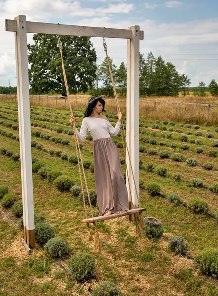 stock image Beautiful young brunette caucasian woman swinging on a wooden swing in a wreath against the lavender field.