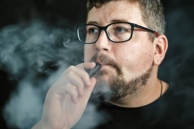 Elektronik sigara içen, gözlük ve küpe takan, kızıl saçlı, sakallı ve siyah arka planda bıyıklı beyaz bir adamın stüdyo portresi..