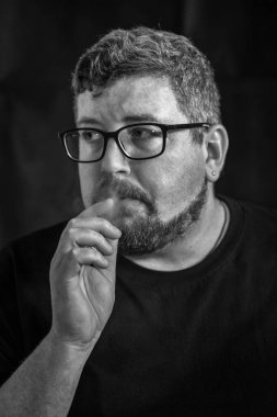 Elektronik sigara içen, siyah arka planda sakal ve bıyıklı, gözlük ve küpe takan beyaz bir adamın stüdyo portresi..