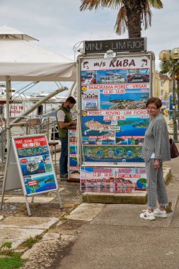 Porec, Hırvatistan - 21 Mayıs 2025: Rovinj, Lim Fjord ve Vrsar 'a tekne gezilerini tanıtan turne standı. Bir kadın, turne seçeneklerini, saatlerini ve fiyatlarını kıyı ortamında gösteren bilgi panolarına bakar