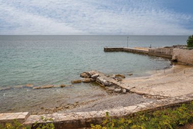 Taş iskele, kayalık plaj ve bulutlu gökyüzünün altında berrak turkuaz deniz. Sakin deniz manzarası, boş kıyı şeridi. Akdeniz yaz havası, seyahat ve tatil konsepti. Istria, Hırvatistan.
