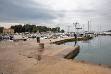 Porec, Hırvatistan - 21 Mayıs 2025: Bulutlu bir günde yat, tekne ve liman manzarası. Sahil sahnesi su, park etmiş araçlar ve çevreleyen yeşillikler üzerine yansıyor..