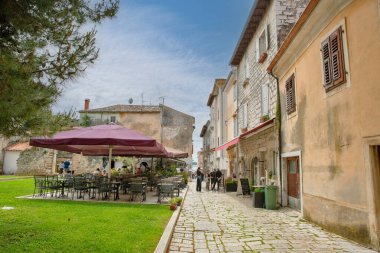 Porec, Hırvatistan - 21 Mayıs 2025: Geleneksel taş binalar, büyük bir şemsiye altında açık bir kafe ve kaldırım taşı patika boyunca yürüyen insanlar ile eski kentte büyüleyici sokak manzarası.
