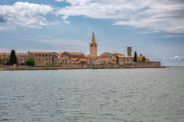 Adriyatik Denizi 'nden Porec' in tarihi şehir yarımadası, Hırvatistan 'da dramatik bulutlu gökyüzü altında ikonik Euphrasian bazilikası, ortaçağ taş binaları ve antik duvarları gösteren çan kulesini gösteriyor.