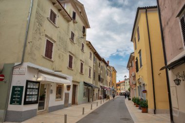 Novigrad, Hırvatistan - 22 Mayıs 2025: Renkli tarihi binalar, yerel dükkanlar ve bulutlu bir gökyüzü altında gezinen insanlar ile eski kentteki huzur dolu yaya caddesi.