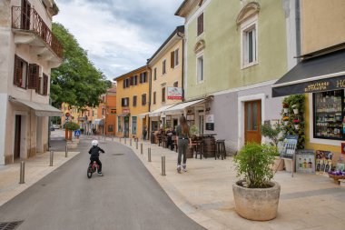 Novigrad, Hırvatistan - 22 Mayıs 2025: Renkli Akdeniz binaları, açık kafeler, dükkanlar ve arabaların keyfini çıkaran turistlerle yaya caddesinde bisiklet süren çocuk.