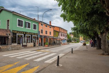 Novigrad, Hırvatistan - 22 Mayıs 2025: Canlı cepheler ve yerel dükkanlar sakin bir caddeyi hizaya getiriyor. Ağaçlarla gölgelendirilmiş kaldırım ve yaya geçidi bu rahat şehir ortamına cazibe katıyor..