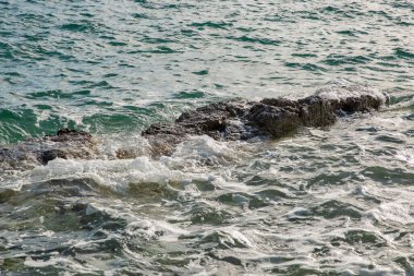 Dinamik deniz manzarası, kıyı şeridi boyunca turkuaz yeşil okyanus suyuyla kırılan ve sıçrayan siyah kayalara köpüklü beyaz dalgalar gösteriyor..