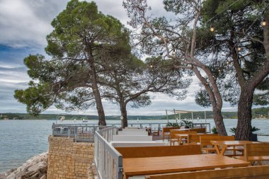 Akdeniz çam ağaçlarının altında ahşap masalar ve banklar bulunan, taş duvarlar, metal korkuluklar ve panoramik su manzaralı güzel bir sahil restoranı terası. Istria, Hırvatistan.