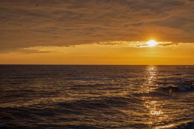 Sakin sulara sıcak turuncu ve sarı renkler yansıtan dramatik bulutlu gökyüzü ile muhteşem bir altın saat deniz manzarası. İskele yapısı barışçıl kıyı atmosferi ekler..