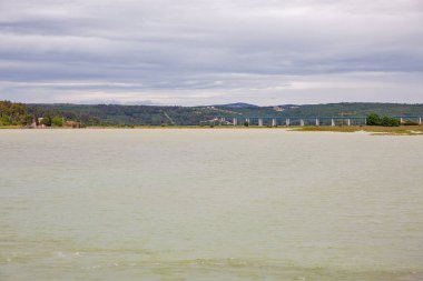 Mirna Köprüsü, arka planda bulutlu gökyüzü ve ormanlık tepelerle huzurlu göl sularını geçerek uzun beton bir yol. Istria Yarımadası, Hırvatistan