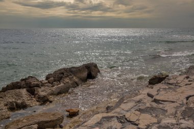 Kasvetli bulutlu gökyüzünün altında kayalara çarpan dalgalarla engebeli taşlı kıyı şeridi. Köpüklü su yansımaları güzel doğal deniz manzarası bileşimi yaratır..