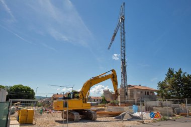 Sarı kazıcı ve kule vinci açık mavi gökyüzünün altındaki bir inşaat sahasında çalışıyor. Endüstriyel ekipman ve inşaat malzemeleri.
