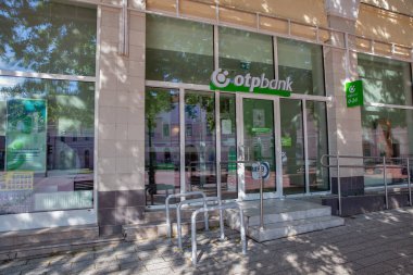 Nagykanizsa, Macaristan - 18 Mayıs 2025: Geniş cam pencereler, yeşil şirket markalaşması ve kentsel caddede benekli güneş ışığı olan metal bisiklet rafları içeren çağdaş OTP Bank şubesi.