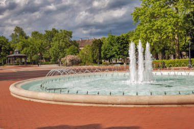 Çevresel bir fıskiyeyi peyzajlı bir şehir parkında birden fazla su püskürtüsü ve püskürtüsüyle süslemek. Kırmızı tuğla plaza, yemyeşil ağaçlar ve tarihi binalarla çeşmeyi çevrelemektedir. Nagykanizsa, Macaristan.