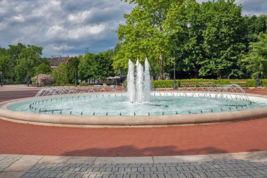 Çevresel bir fıskiyeyi peyzajlı bir şehir parkında birden fazla su püskürtüsü ve püskürtüsüyle süslemek. Kırmızı tuğla plaza, yemyeşil ağaçlar ve tarihi binalarla çeşmeyi çevrelemektedir. Nagykanizsa, Macaristan.