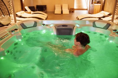 Orta yaşlı bir kadın, sıcak, aydınlık bir jakuzide, yumuşak bir ışık ve sakin bir atmosferle çevrili, huzurlu bir şekilde banyo yapıyor..