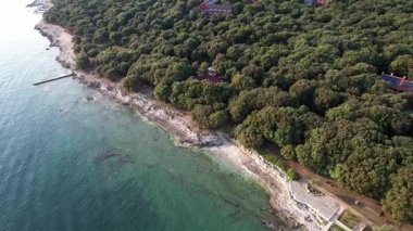 Hırvatistan 'ın Istria kentindeki kayalık bir günbatımı sahil şeridi olan Lanterna yarımadasının hava aracı görüntüsü, kristal berrak turkuaz su, sörf dalgaları, kıyıya taş basamaklar ve uçurumların üzerindeki yemyeşil orman.