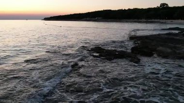 Gün batımında yumuşak deniz dalgaları pastel gökyüzü ve ağaçların siluetiyle kayalık kıyılara vurur. Huzurlu sahil manzarası seyahat, doğa ve rahatlama temaları için mükemmel. Istria, Hırvatistan.