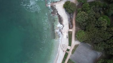 Hırvatistan 'ın Istria kentindeki Lanterna yarımadasındaki kayalık bir sahil sahil şeridinin hava aracı görüntüsü, kristal berrak turkuaz su, sörf dalgaları ve kayalıkların üzerindeki yemyeşil ormanlarla dolu..