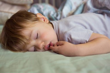 Tatlı uyuyan bebek. Çocukların gündüz uykusu. Küçük beyaz çocuk büyük bir yatakta, battaniyenin altında uyuyor. 1-2 yaşında uyuyan güzel sarışın bir çocuğun yakın plan portresi.. 