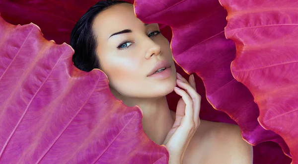 Young woman\'s face surrounded by magenta tropical leaves. Woman face with Natural make-up on a tropical leaf pink background. Natural cosmetic and summer wellness. Purity and skincare. Space for text