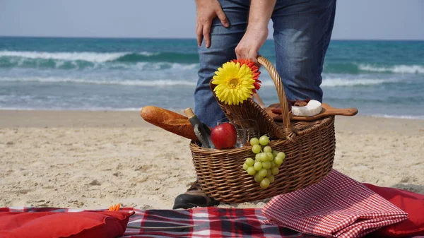 A man holds a wicker basket with food and wine for picnic on the beach. Summertime relaxation and recreation concept.