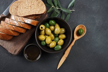 Renkli zeytinler, zeytin dalı, kapta zeytin yağı, arka planda dilimlenmiş ekmek.