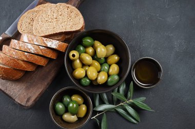 Renkli zeytinler, zeytin dalı, kapta zeytin yağı, arka planda dilimlenmiş ekmek.