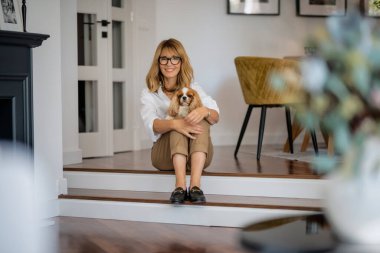 Orta yaşlı, mutlu bir kadın yerde beyaz şirin köpeğiyle oturuyor. Evde dinlenirken gözlük takan çekici bir kadın..